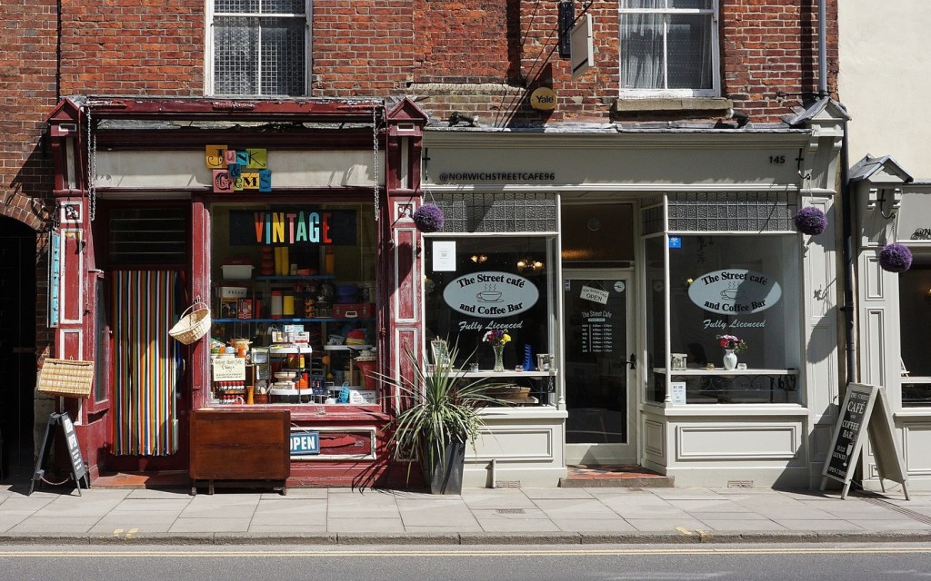 Two shops on a street, one a cafe and the other selling vintage goods. The building is made of brick and it's sunny outside.