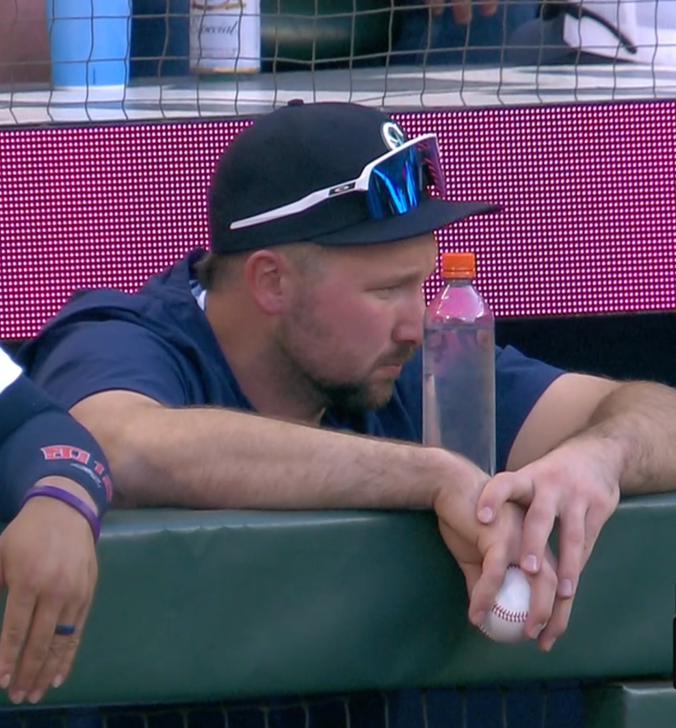 Cal Raleigh leaning against the dugout railing while holding a baseball (transitional object) in his right hand. A water bottle is directly in front of his face on the railing. 